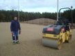 John Sildeøje er steget ned fra bulldozeren, og kører med tromlen. I baggrunde "Sildeøje Bakken", som John har skabt til glæde for alle børn, der kan lide at kælke.
Underlaget for kunstgræsset er nu 100% afrettet, og der kørers med tromlen for at gøre underlaget helt klart til kunstgræsset. For at gøre det helt lige afsluttes med en lille manuel håndtromle, der fuldender finish. Herefter er det klart til at der skal rulles kunstgræs ud.